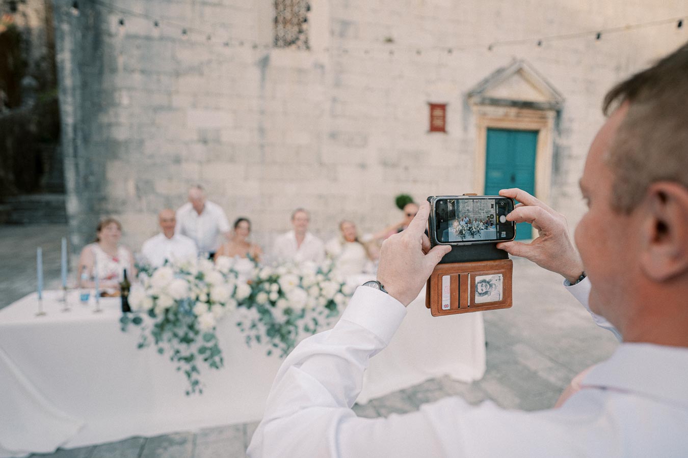 A person taking a photo of a wedding party seated at a decorated table outside, with a smartphone in a leather case, against a historic stone building backdrop.