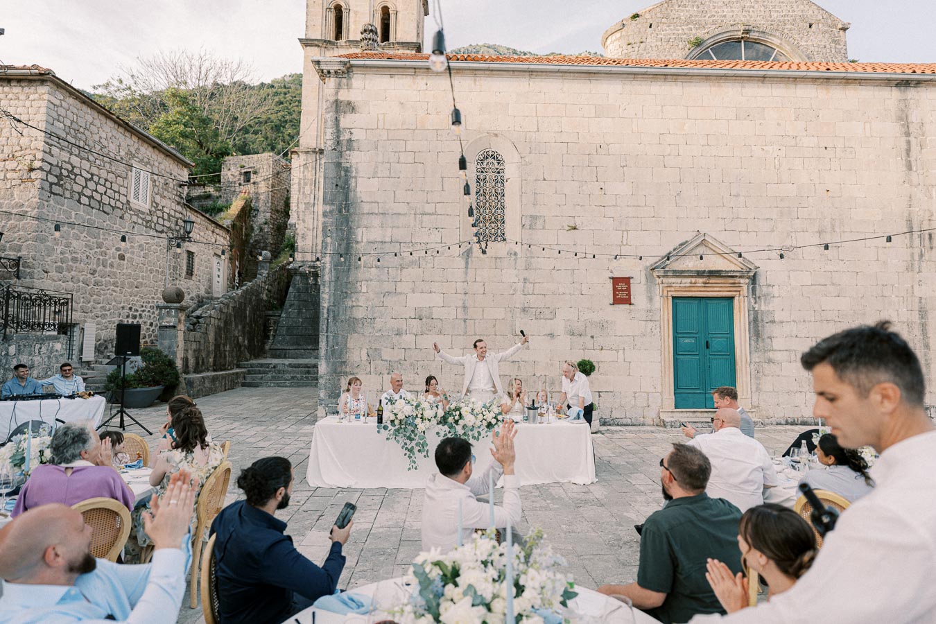 Outdoor wedding reception with guests seated around tables, a speaker standing with a microphone in front of a historic stone building adorned with string lights and floral decorations.