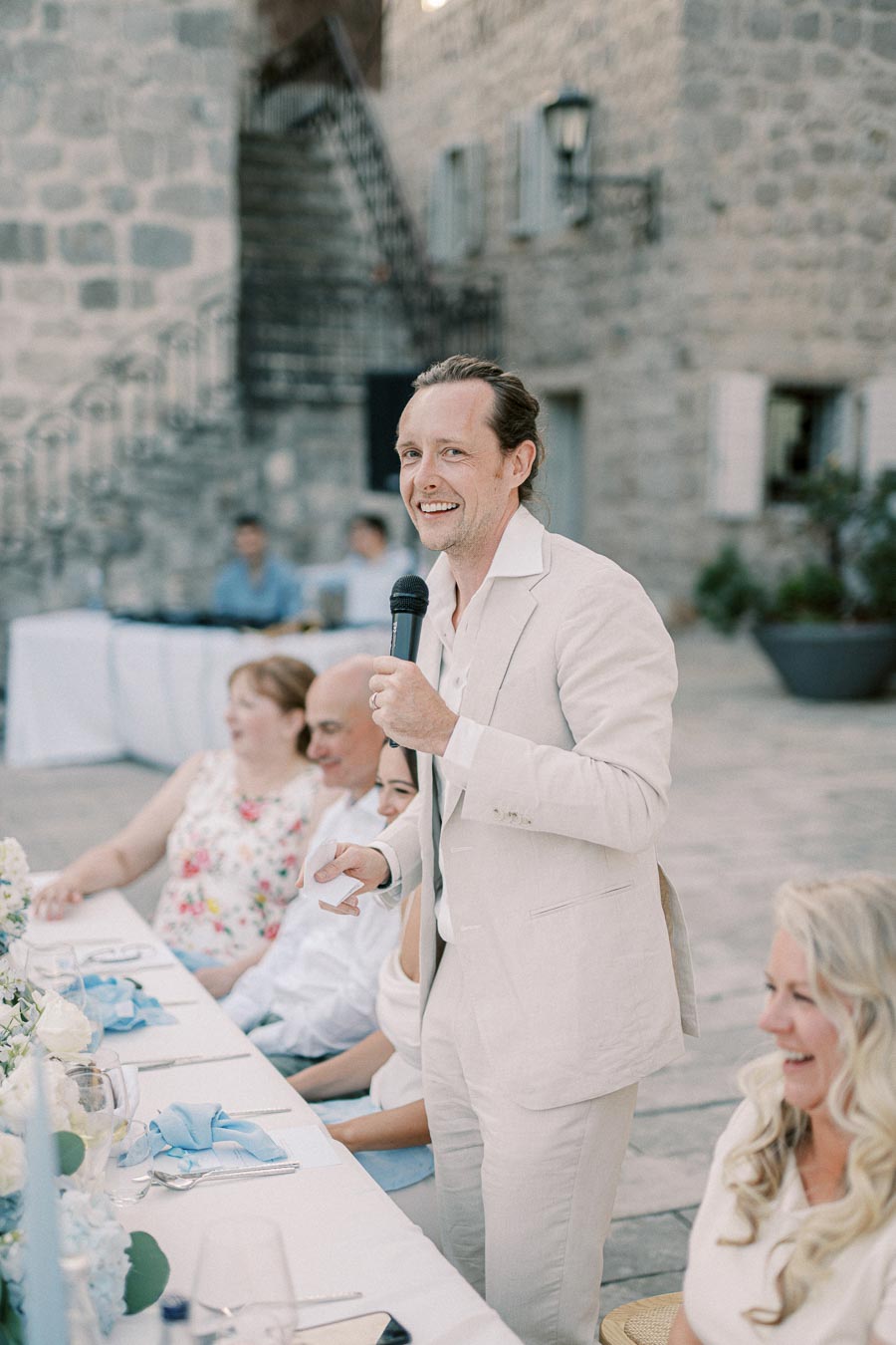 A man in a light suit giving a speech with a microphone at an outdoor event, standing next to a table with seated guests and floral decorations. Stone walls and stairs in the background create a rustic atmosphere.