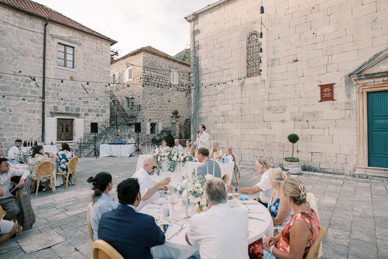 Elegant outdoor wedding reception at a historic stone courtyard with guests seated at round tables decorated with floral arrangements, under string lights.