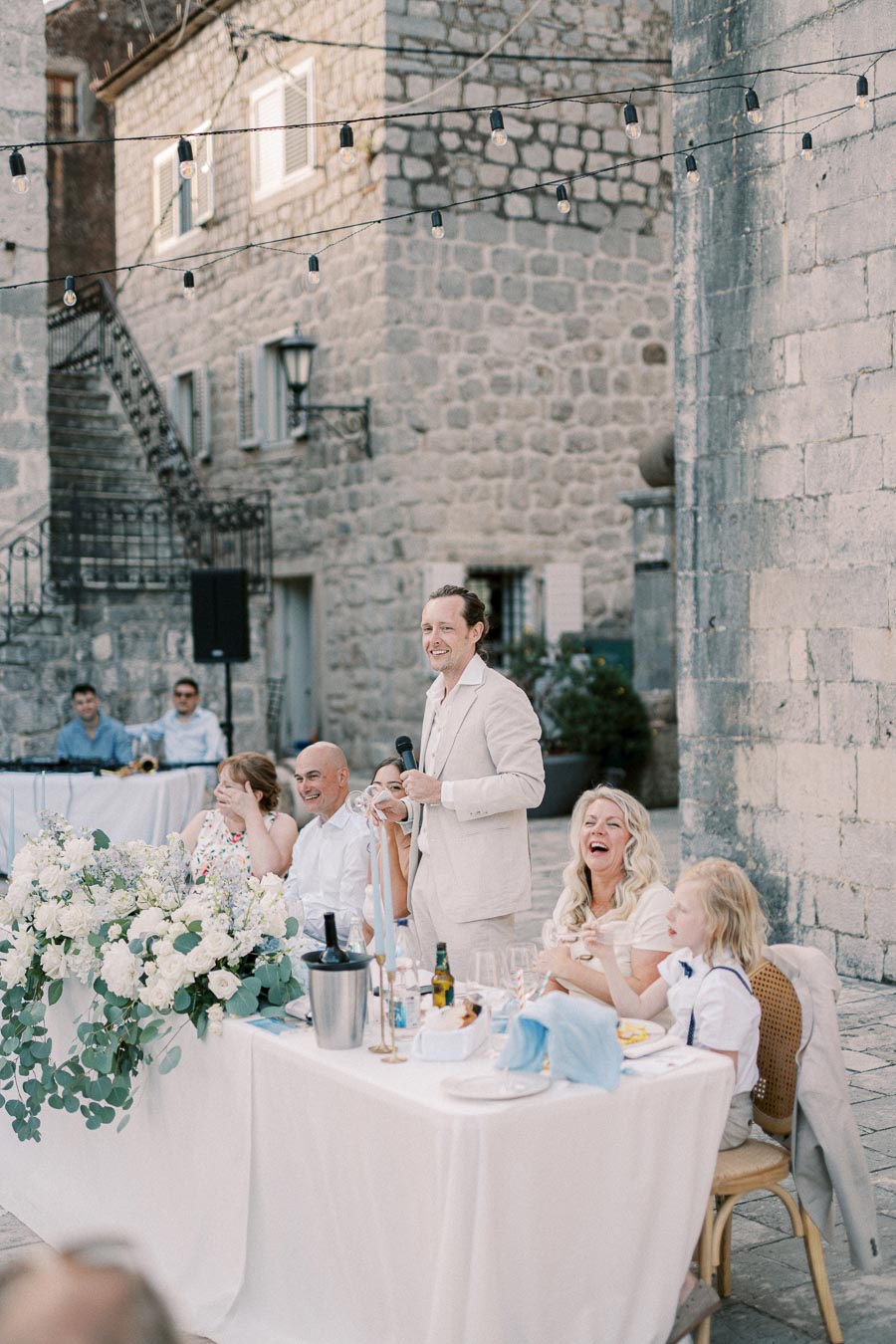 Elegant outdoor wedding reception with guests enjoying a speech by a man in a beige suit, surrounded by rustic stone architecture and string lights, creating a romantic atmosphere.