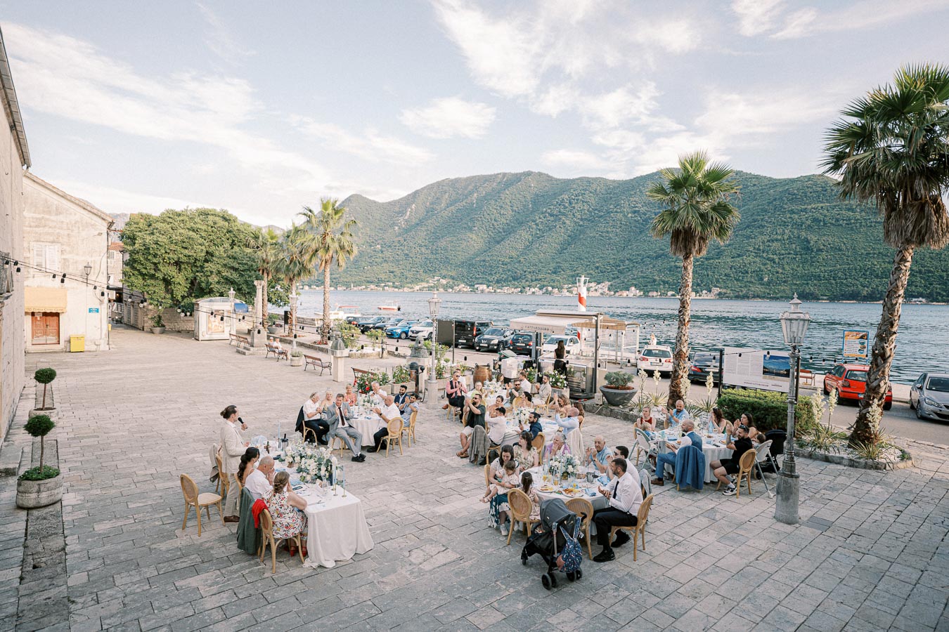 Outdoor wedding reception by the sea with guests seated at elegantly decorated tables under a bright blue sky, surrounded by palm trees and scenic mountain views in the background.