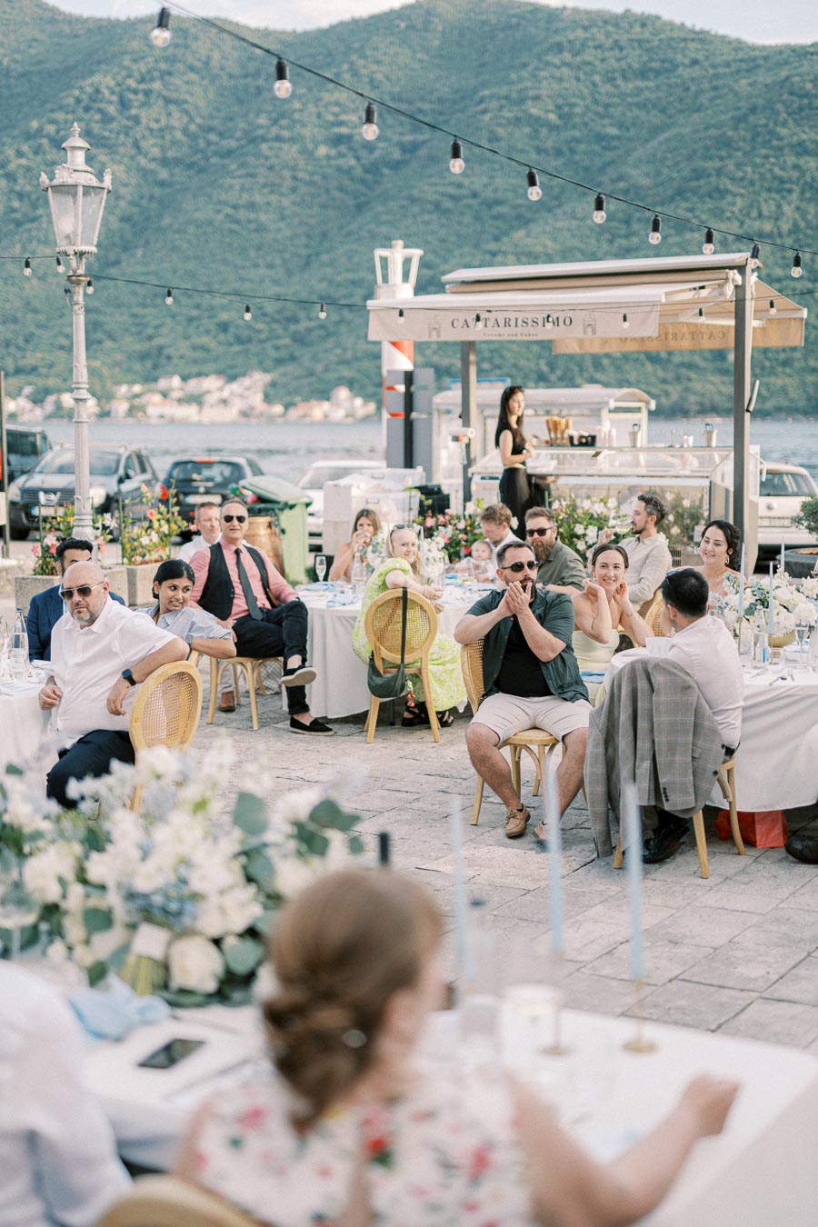 Outdoor wedding reception by the lake, featuring guests seated at decorated tables under string lights, with scenic mountain views and a casual, festive atmosphere.