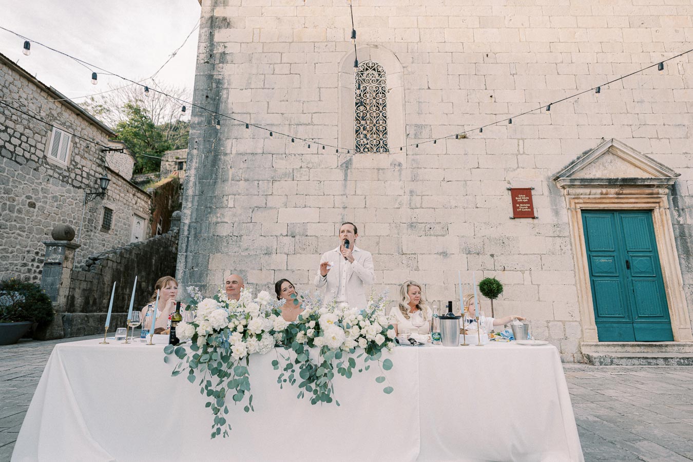 Outdoor wedding reception with a speaker at a table decorated with lush floral arrangements. Stone building and teal door in the background.