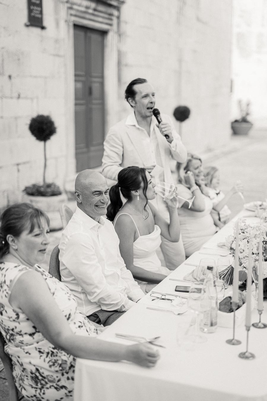 Black and white photo of a wedding reception with guests seated at a long table. A man stands and speaks into a microphone, while other guests smile and listen in a relaxed, outdoor setting.