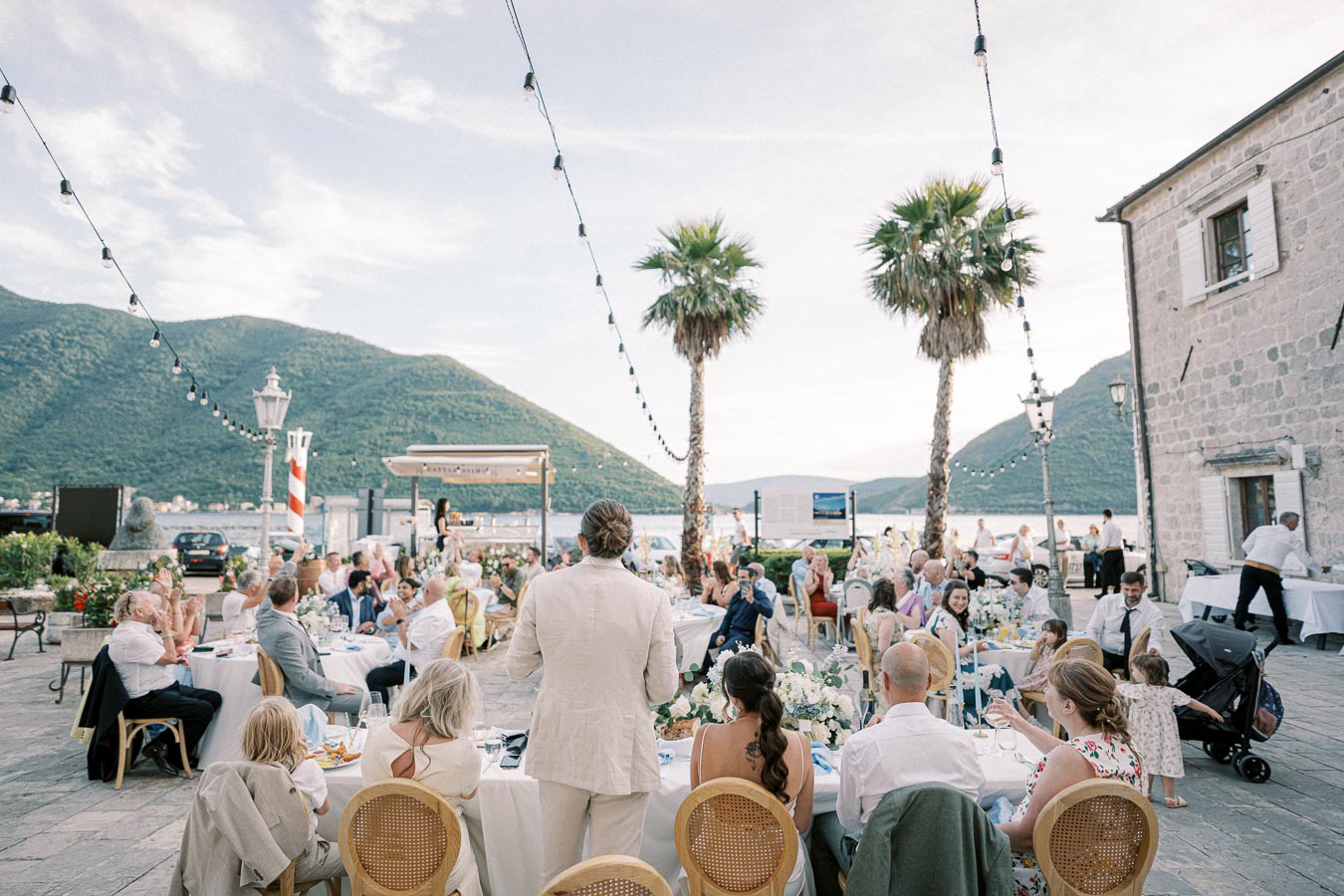 Outdoor wedding reception by a scenic waterfront with palm trees, overlooking mountains, featuring elegantly dressed guests seated at decorated tables during sunset.