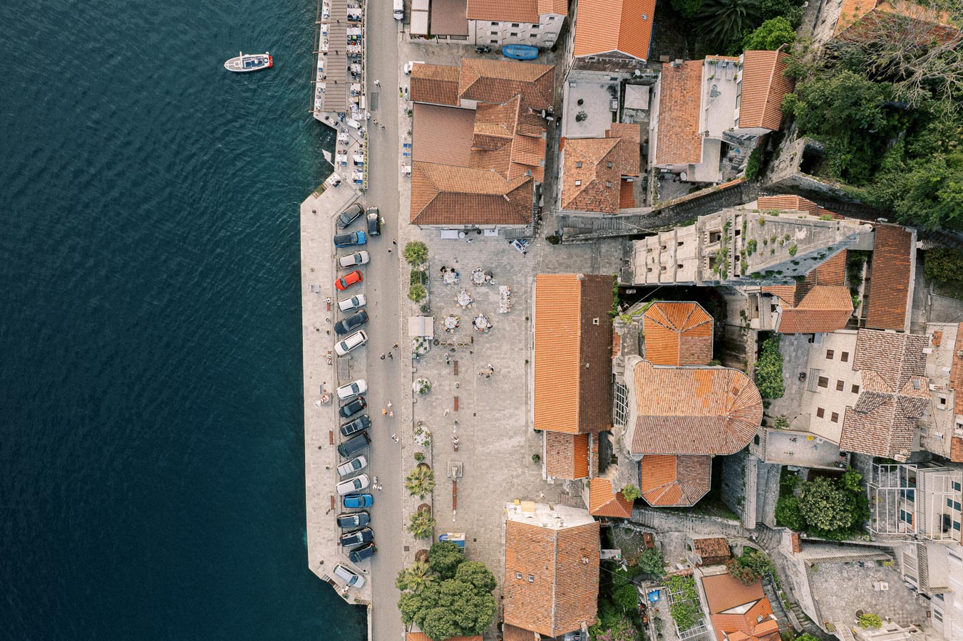 Aerial view of a coastal town with red-tiled rooftops, a docked boat, cars parked along the waterfront, and outdoor seating areas, located by the sea with vivid blue water.