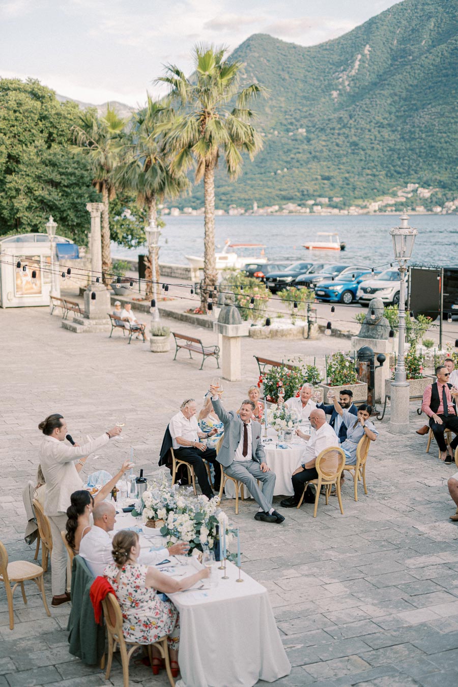 A waterfront wedding reception with guests raising their glasses for a toast in an outdoor setting, featuring elegant white tables and floral centerpieces, with a picturesque backdrop of palm trees and scenic mountains.