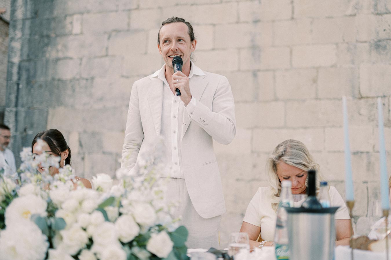 Man in a white suit giving a speech with a microphone at an outdoor wedding ceremony, surrounded by floral decorations and seated guests.
