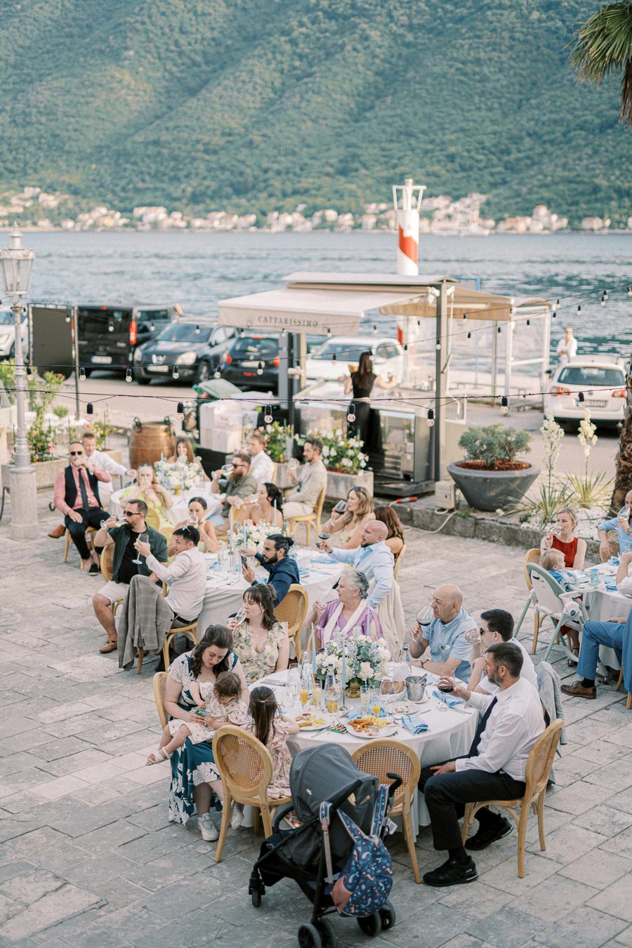 A group of people enjoying an outdoor celebration by a lakeside with scenic mountain views in the background, tables elegantly decorated with flowers and drinks, creating a picturesque event setting.