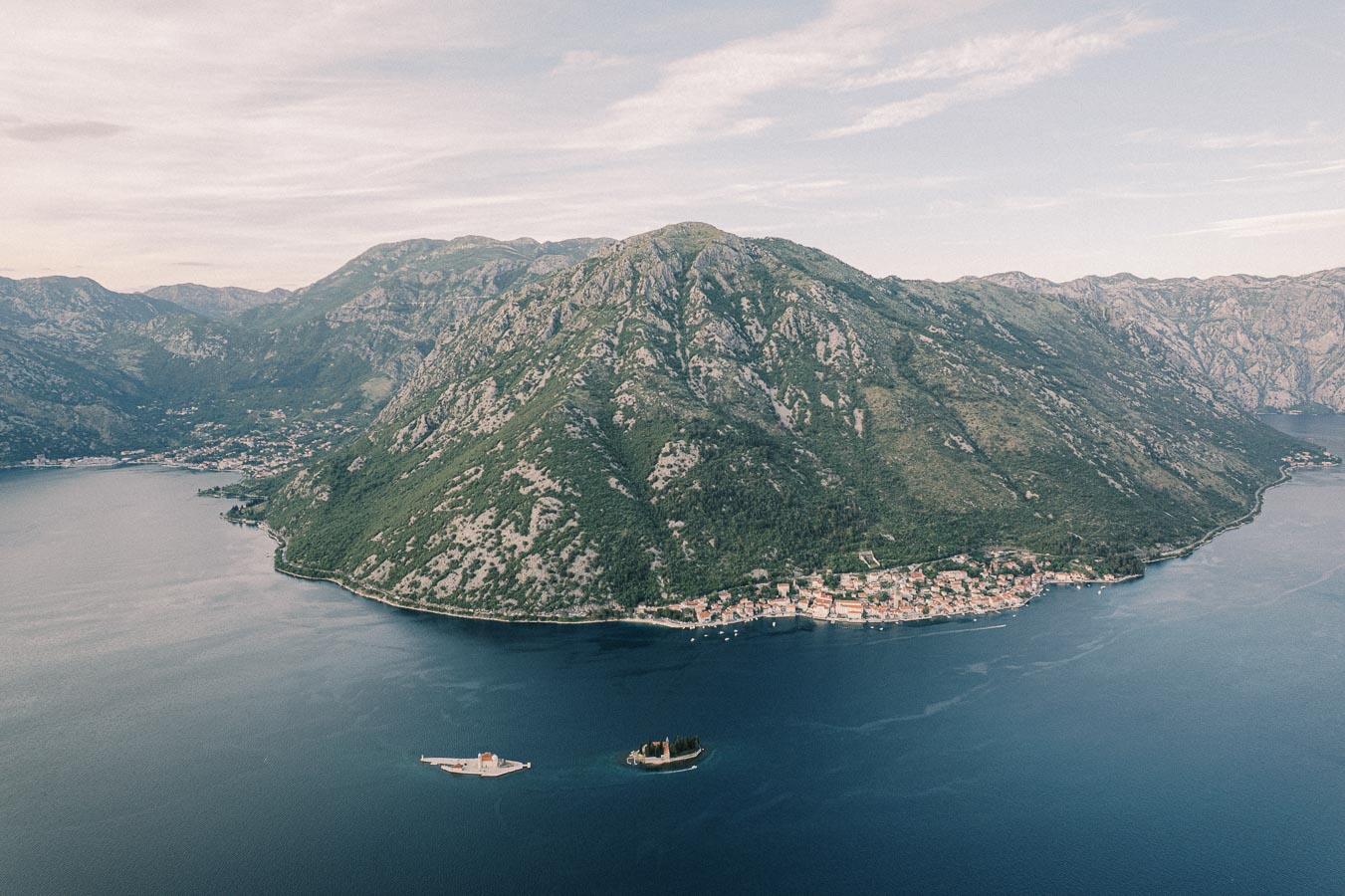 Aerial view of the picturesque Kotor Bay in Montenegro, surrounded by rugged mountains and featuring two small islands. The clear blue water highlights the scenic beauty and tranquil setting of the coastal town, perfect for travel and tourism.
