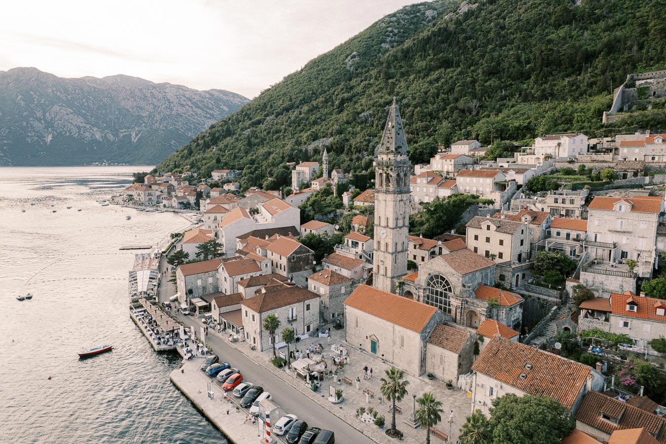 Aerial view of a picturesque coastal town with historic stone buildings and a prominent church tower, nestled between lush green hills and a serene waterfront, showcasing traditional Mediterranean architecture.