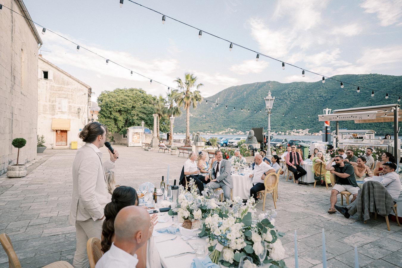 Outdoor wedding reception by the sea with mountains in the background, featuring elegantly dressed guests seated at tables adorned with white flowers, as a man in a light suit gives a speech under string lights.