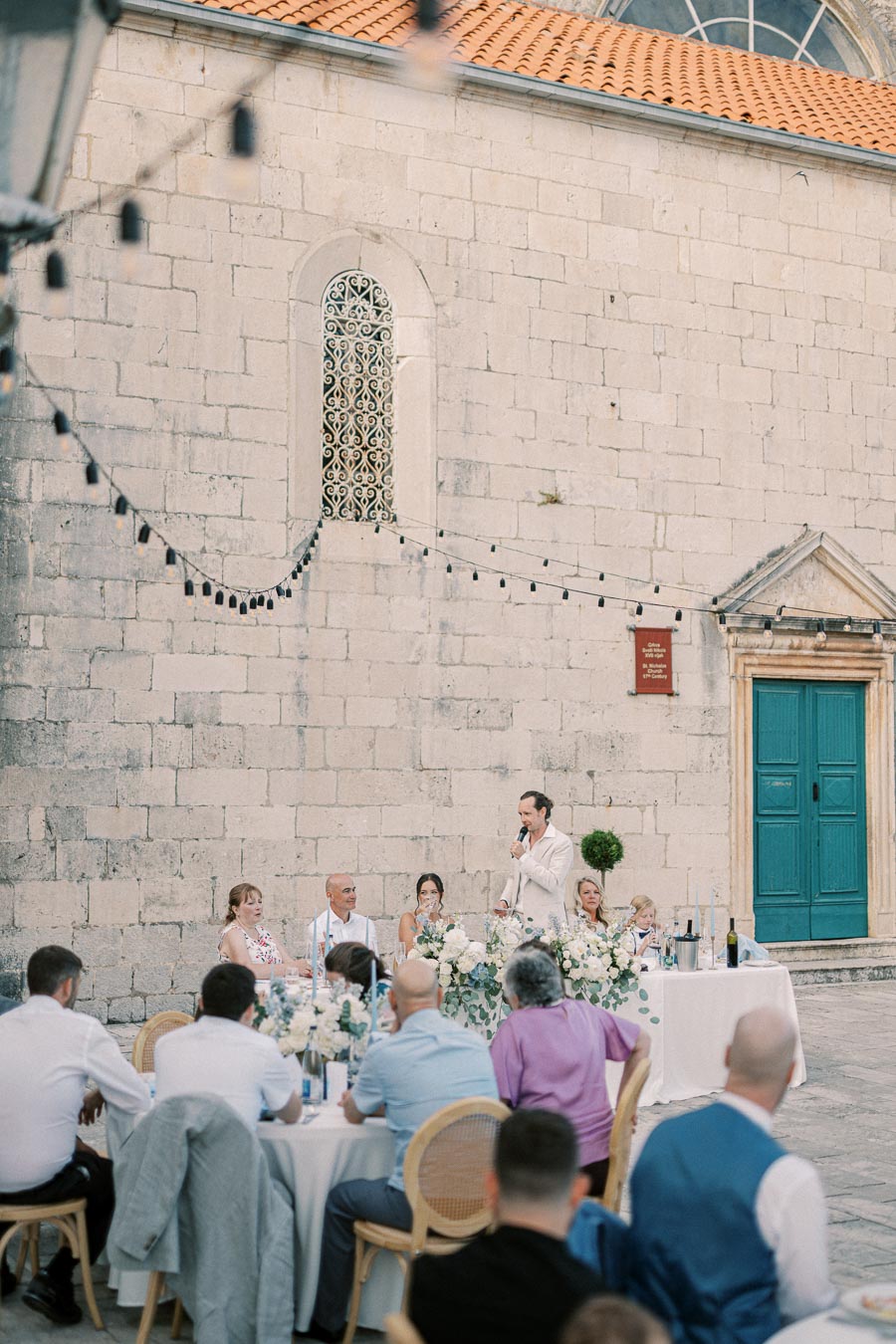 Outdoor wedding reception with guests seated at tables under string lights near a historic stone building, featuring a speaker giving a toast.