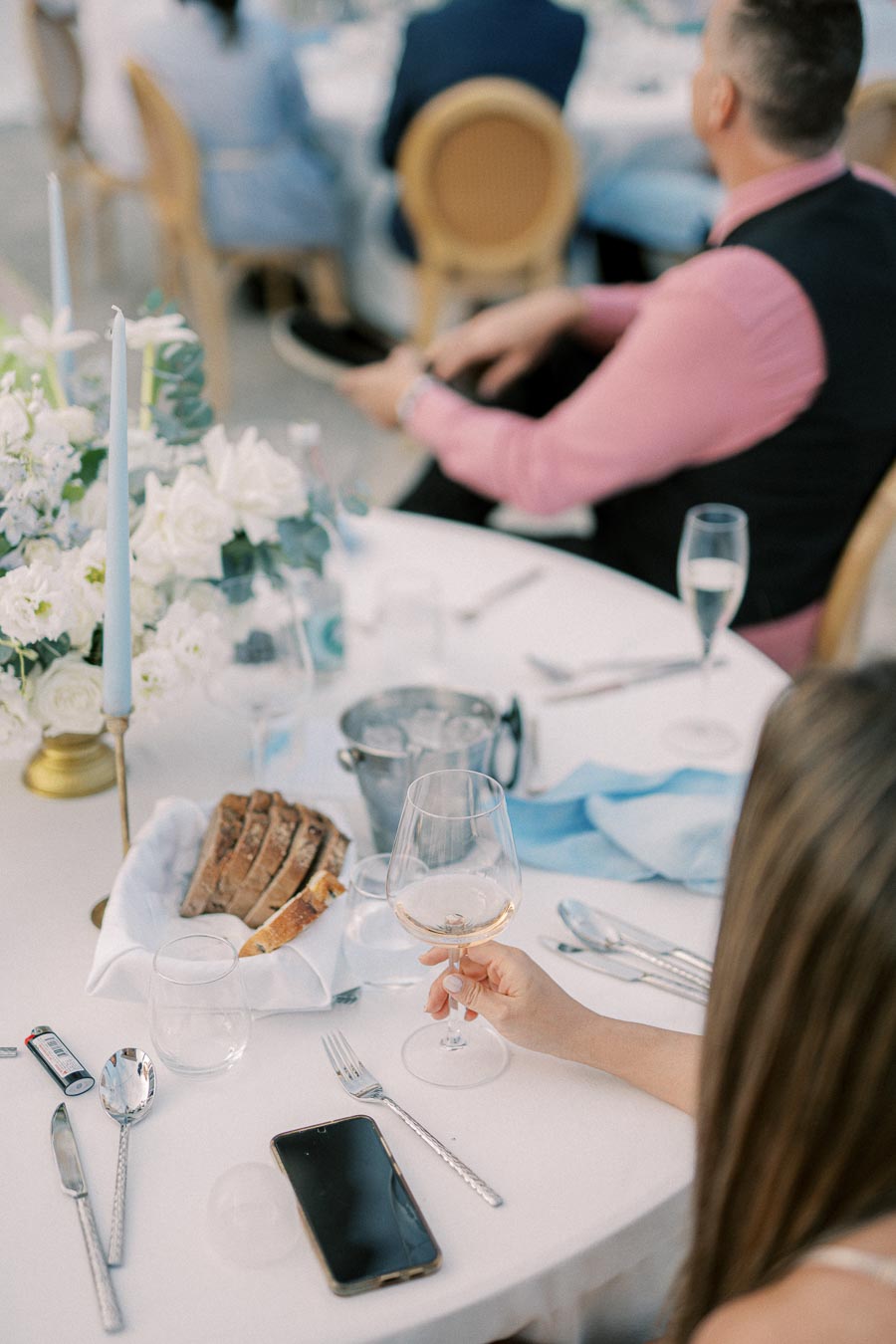 Elegant dinner table setting with a focus on a hand holding a wine glass. The table features a white tablecloth, a basket of sliced bread, cutlery, and a smartphone. In the background, guests are seated at a formal event. The ambiance is enhanced by floral arrangements and a candle.