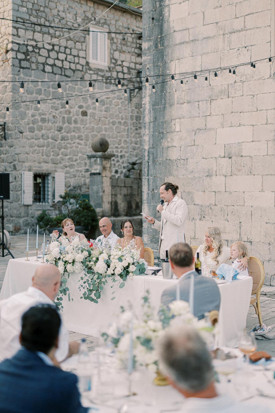 Outdoor wedding reception in a rustic stone courtyard, featuring a speaker addressing guests at a decorated table with floral arrangements and string lights overhead.