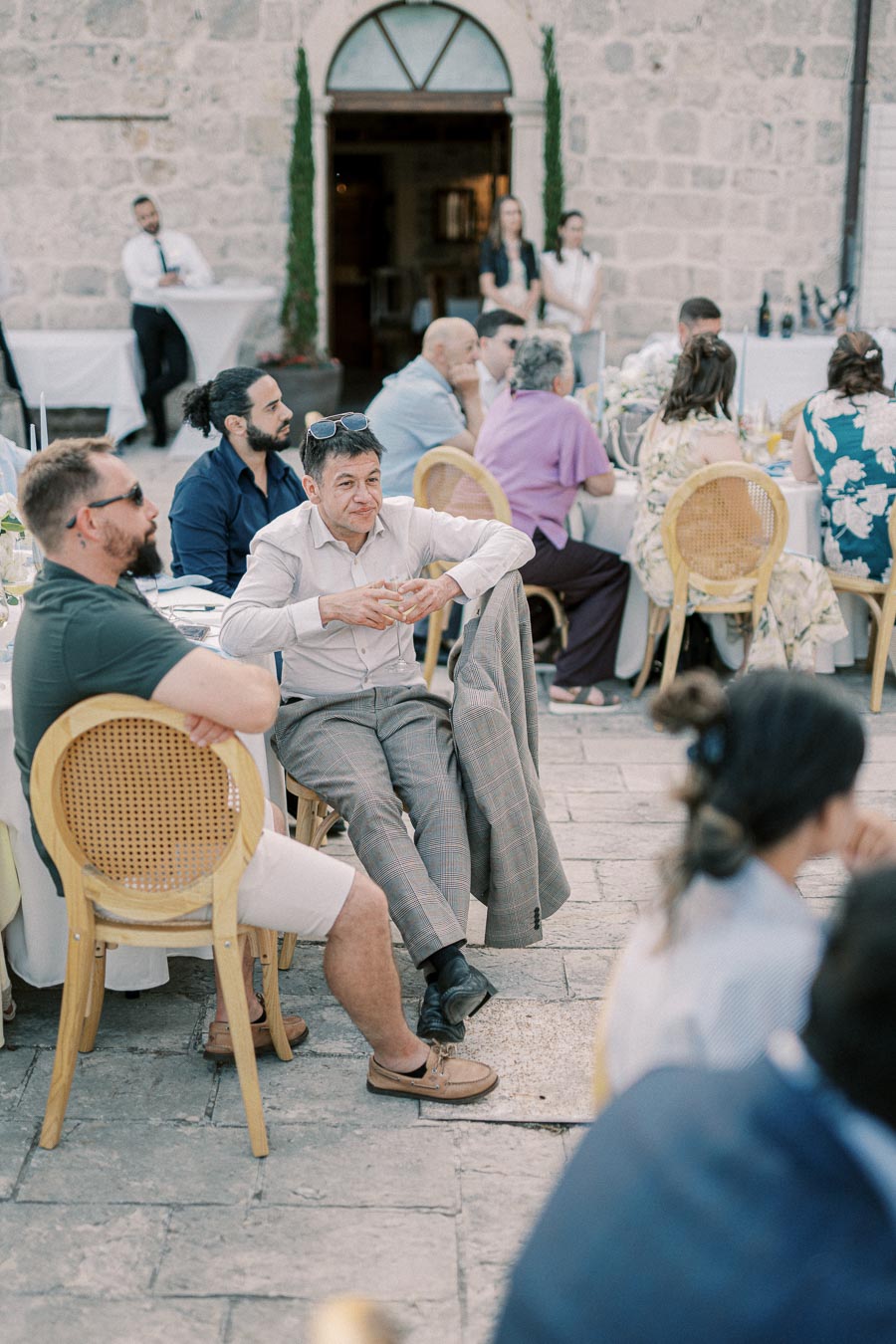 A group of people seated outdoors at a wedding reception, enjoying the event with drinks in hand, surrounded by rustic stone architecture and elegant decorations.