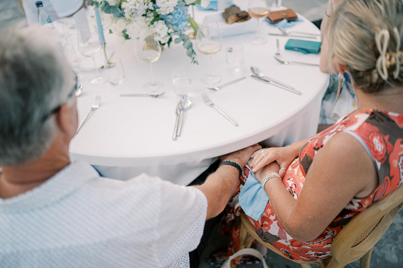 A couple holding hands at an elegantly set table, adorned with white and blue floral centerpieces, creating a romantic and intimate atmosphere at an outdoor event.