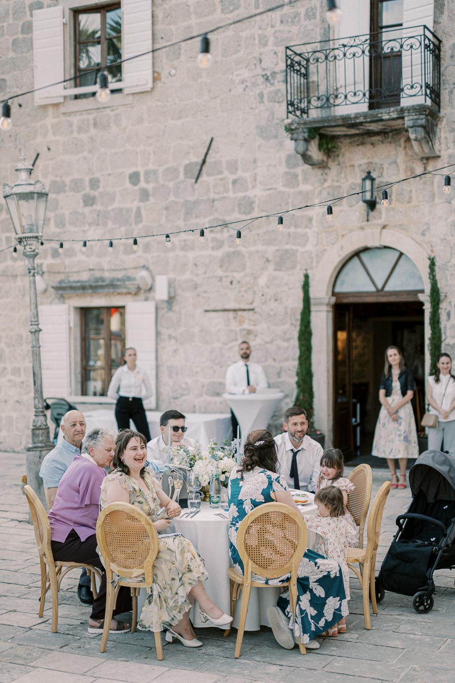 Outdoor wedding reception with guests seated at a decorated table, surrounded by rustic stone architecture and string lights, creating a festive ambiance.
