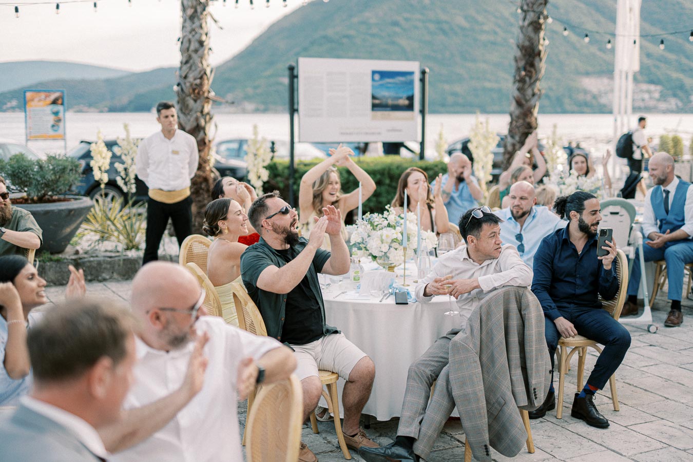 Outdoor event with people sitting at a decorated table, clapping and taking photos, with a scenic mountain and lake view in the background.