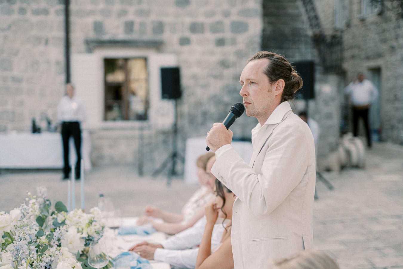 A man in a white suit giving a speech with a microphone at an outdoor event, with floral decorations and blurred guests in the background. Stone wall can be seen, suggesting a rustic venue setting.