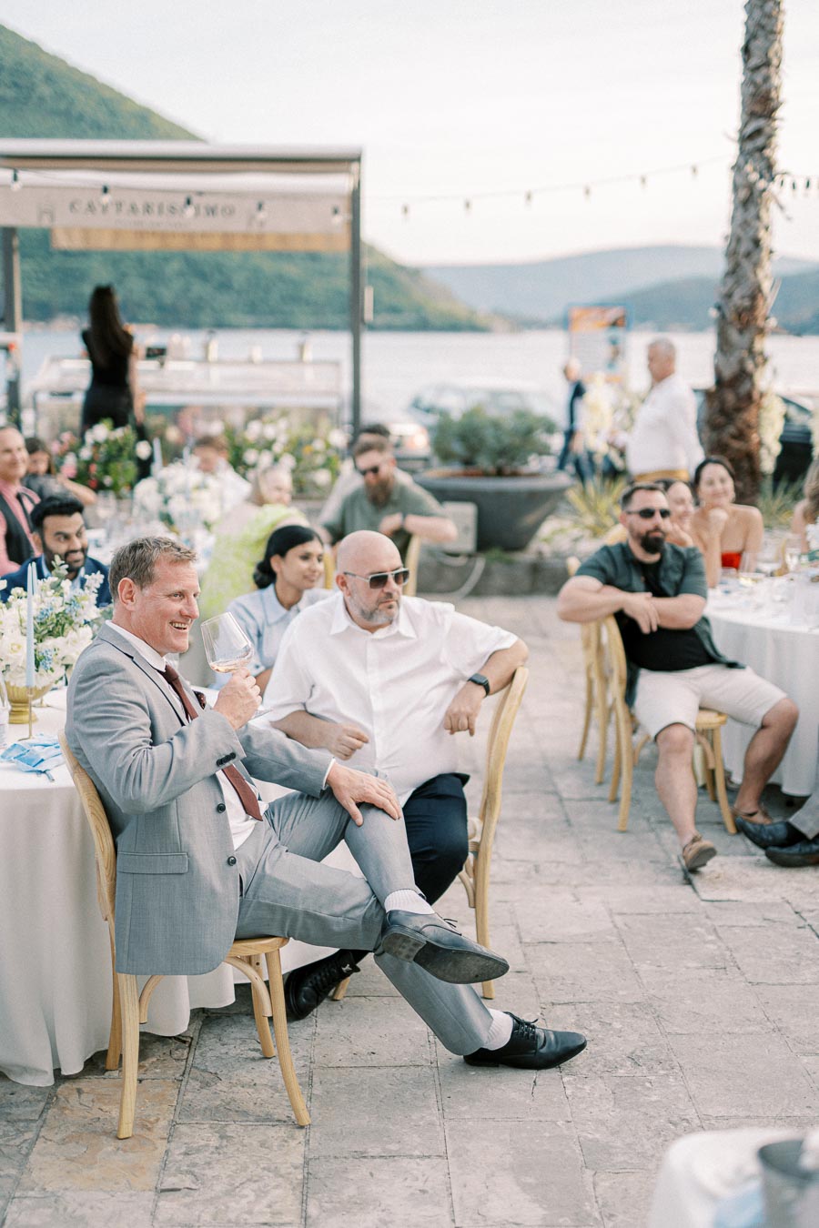 A group of people enjoying an outdoor event by the seaside, featuring elegantly set tables and scenic hill views in the background.