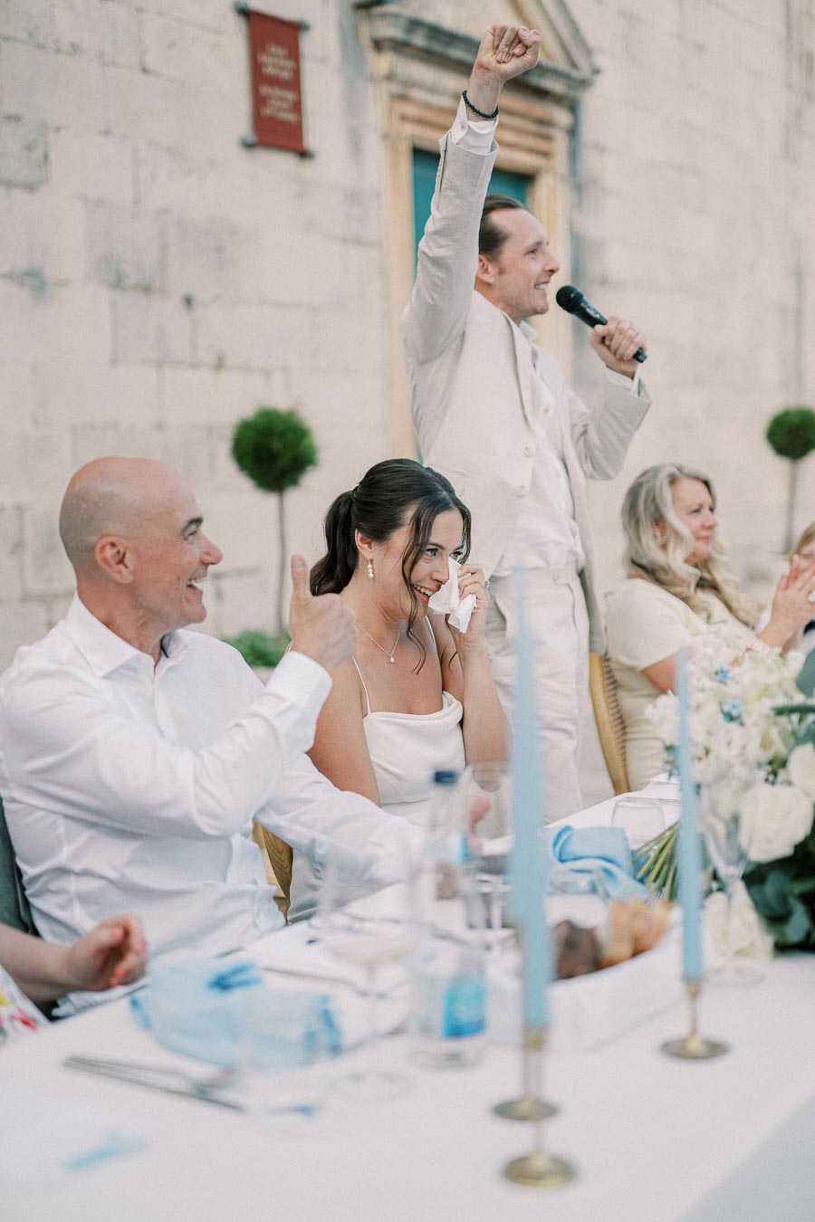 Wedding reception with a joyful speaker holding a microphone, guests giving thumbs up, and a bride in a white dress wiping tears of joy, surrounded by elegant table settings and floral decorations.