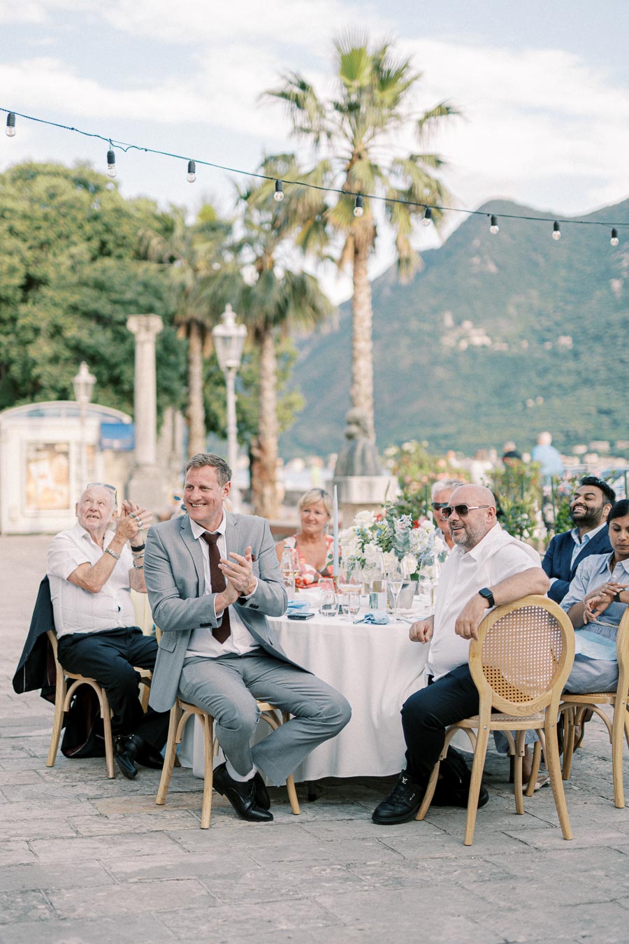 Outdoor wedding reception with guests seated at a round table, featuring decor with white flowers and greenery, palm trees, and mountains in the background, under string lights in a picturesque setting.