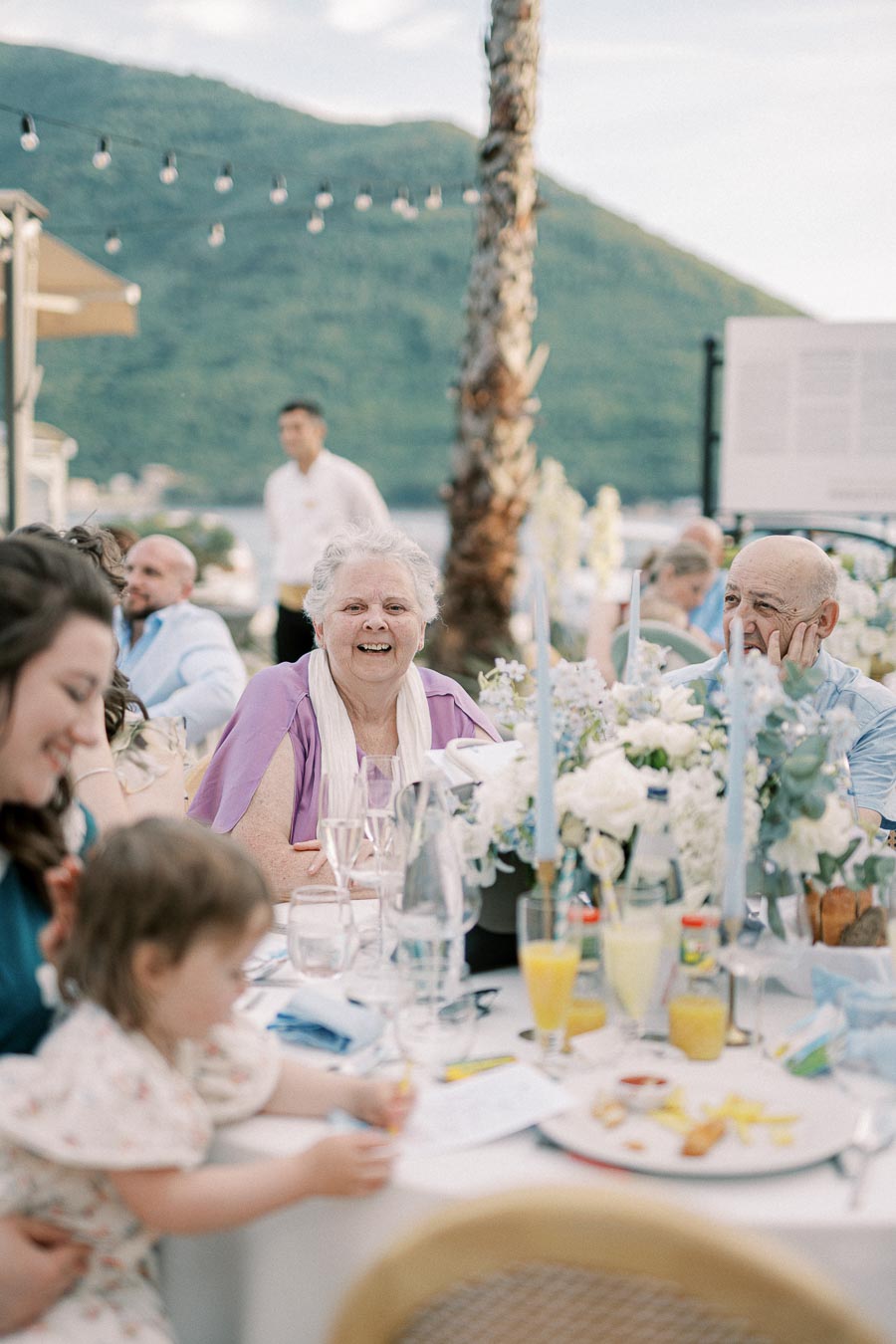 Elderly woman smiling at outdoor family gathering, seated at a table with white and blue floral centerpieces, mountains in the background.