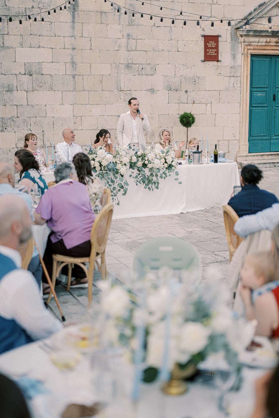 Outdoor wedding reception with a group of people seated at a long table in front of a stone wall, decorated with white flowers and greenery. A man, possibly the host, is speaking into a microphone, while guests enjoy the celebration. Bright string lights are hung above, creating a festive atmosphere.