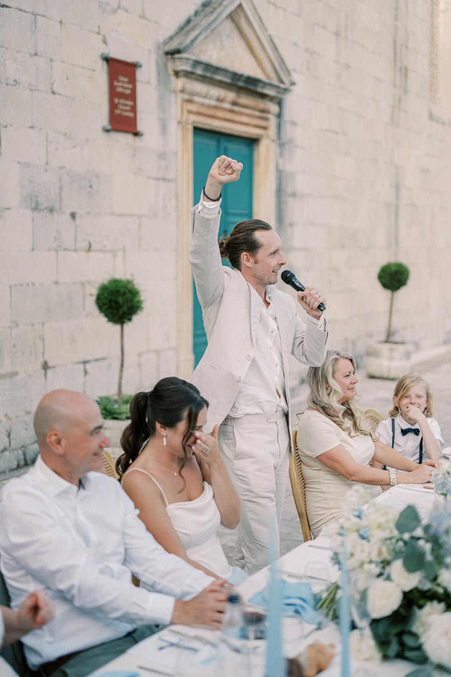 Wedding reception speech outdoors with a man in a beige suit holding a microphone, family seated at a long table with floral decorations, a stone building facade with a blue door in the background.
