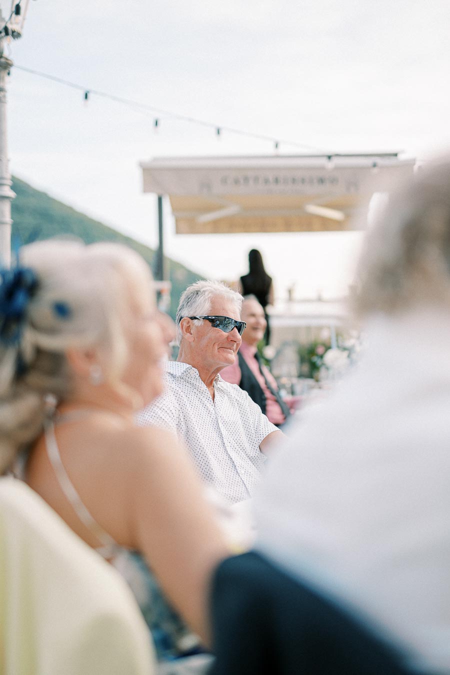 Elderly man wearing sunglasses and a white shirt sitting outdoors at a social event, with blurred guests in the foreground and a mountain landscape in the background.