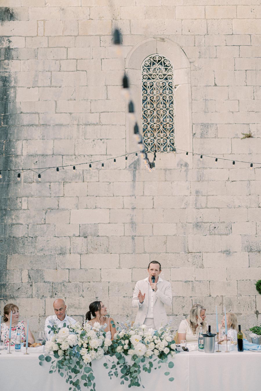 A person giving a speech at an outdoor wedding reception. The head table is adorned with white floral arrangements and greenery, set against a historic stone wall with an ornate window. String lights hang above, enhancing the romantic ambiance.