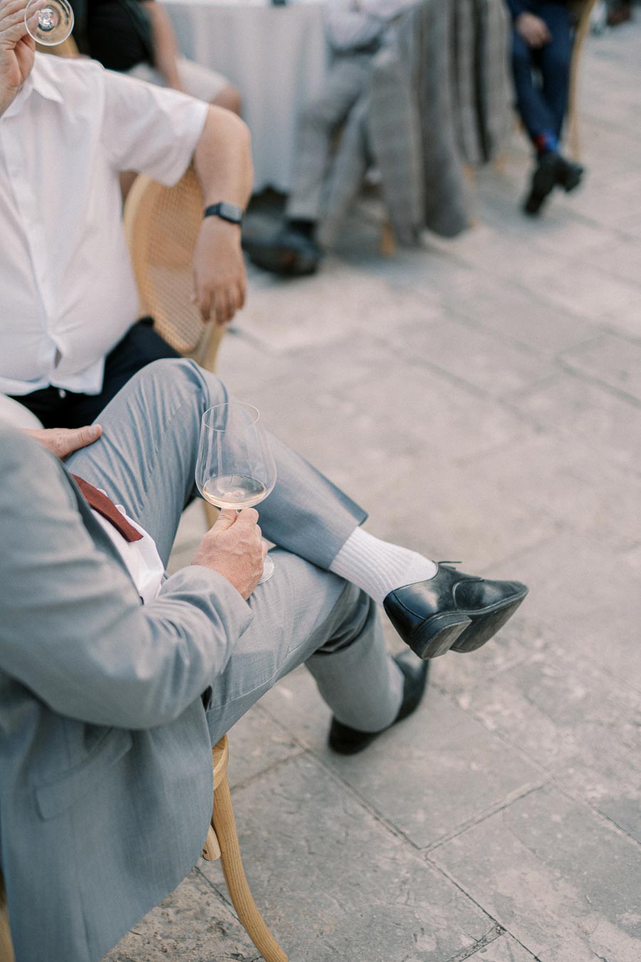 A man in a light gray suit sitting on a wooden chair, holding a glass of white wine outdoors at a social event.