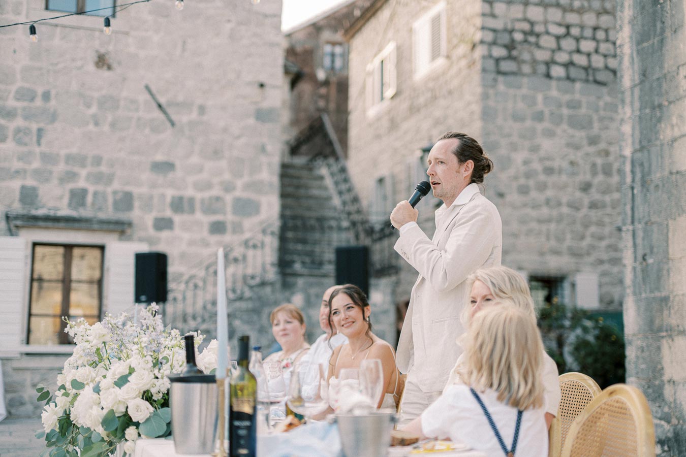 A man giving a speech with a microphone at an outdoor gathering, surrounded by people seated at a long table adorned with elegant white floral arrangements and wine bottles, set against a backdrop of rustic stone buildings.