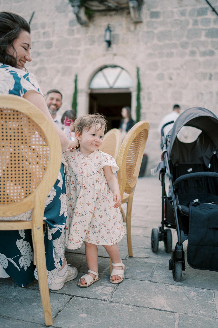 A joyful toddler in a floral dress stands beside an adult seated in a chair outdoors, with a stroller nearby and a stone building in the background, capturing a candid family moment.
