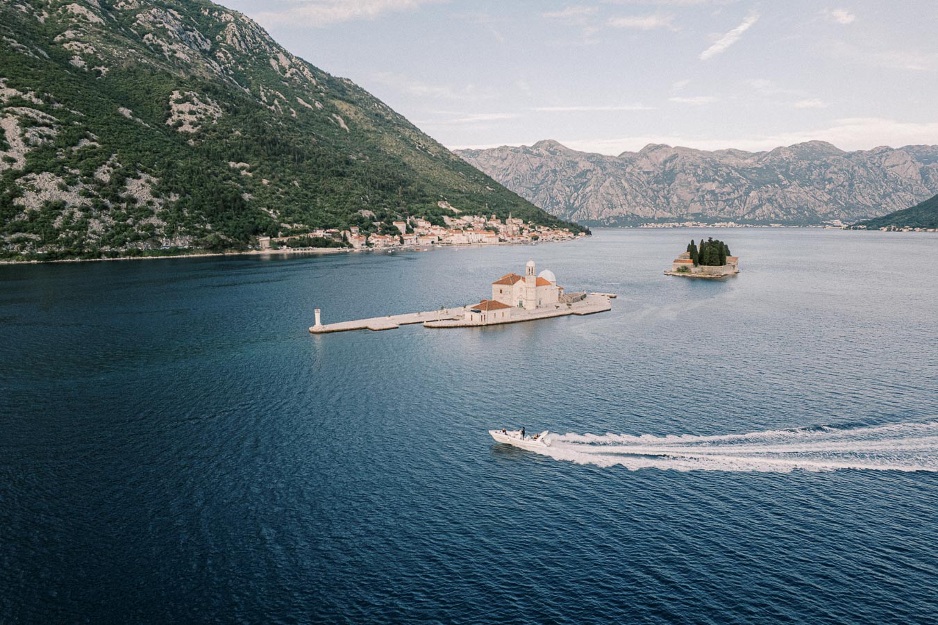 Aerial view of the picturesque Bay of Kotor in Montenegro, featuring two small islands: the Our Lady of the Rocks with its historic church, and the lush, forested Island of St. George. A motorboat zips across the deep blue waters, while the background showcases the coastal town and rugged mountains.