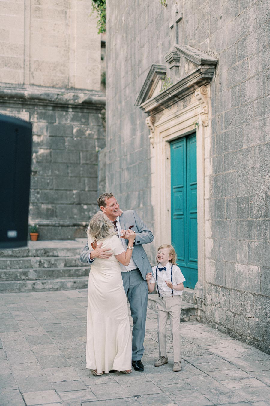 A joyful family moment captured outdoors near an old stone building, featuring a man in a suit, a woman in a white dress, and a smiling child in suspenders and bow tie. The scene conveys happiness and togetherness.