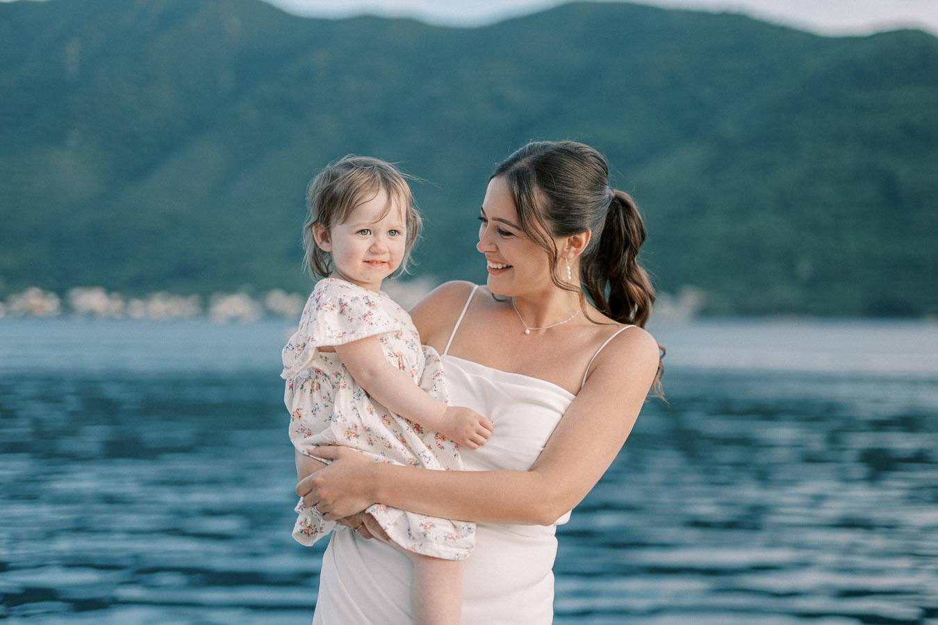 Smiling woman in white dress holding a child near a peaceful lake with mountain backdrop on a sunny day.
