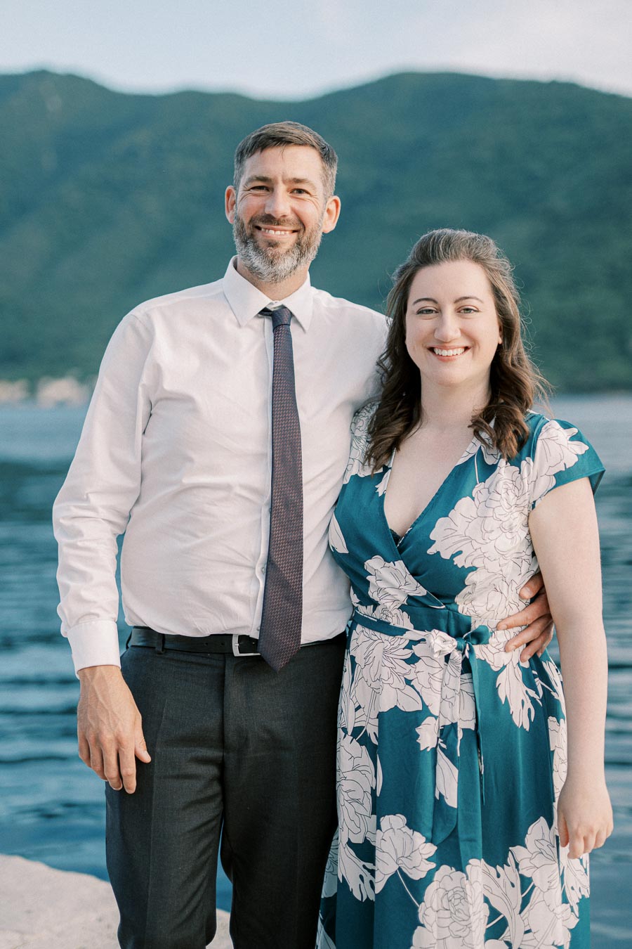 A couple smiling by a scenic lakeside, with lush green mountains in the background. The man is wearing a white shirt and tie, while the woman is in a floral dress, capturing a joyful outdoor moment.