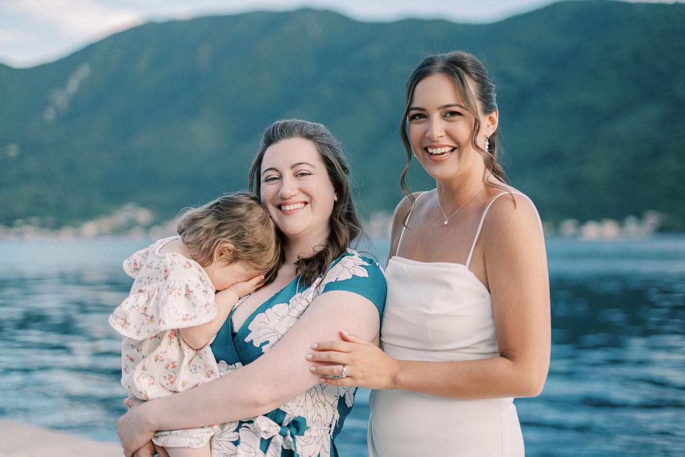 Two smiling women stand by a serene lake, accompanied by a child in a floral dress. The backdrop features lush green mountains, creating a picturesque outdoor scene.