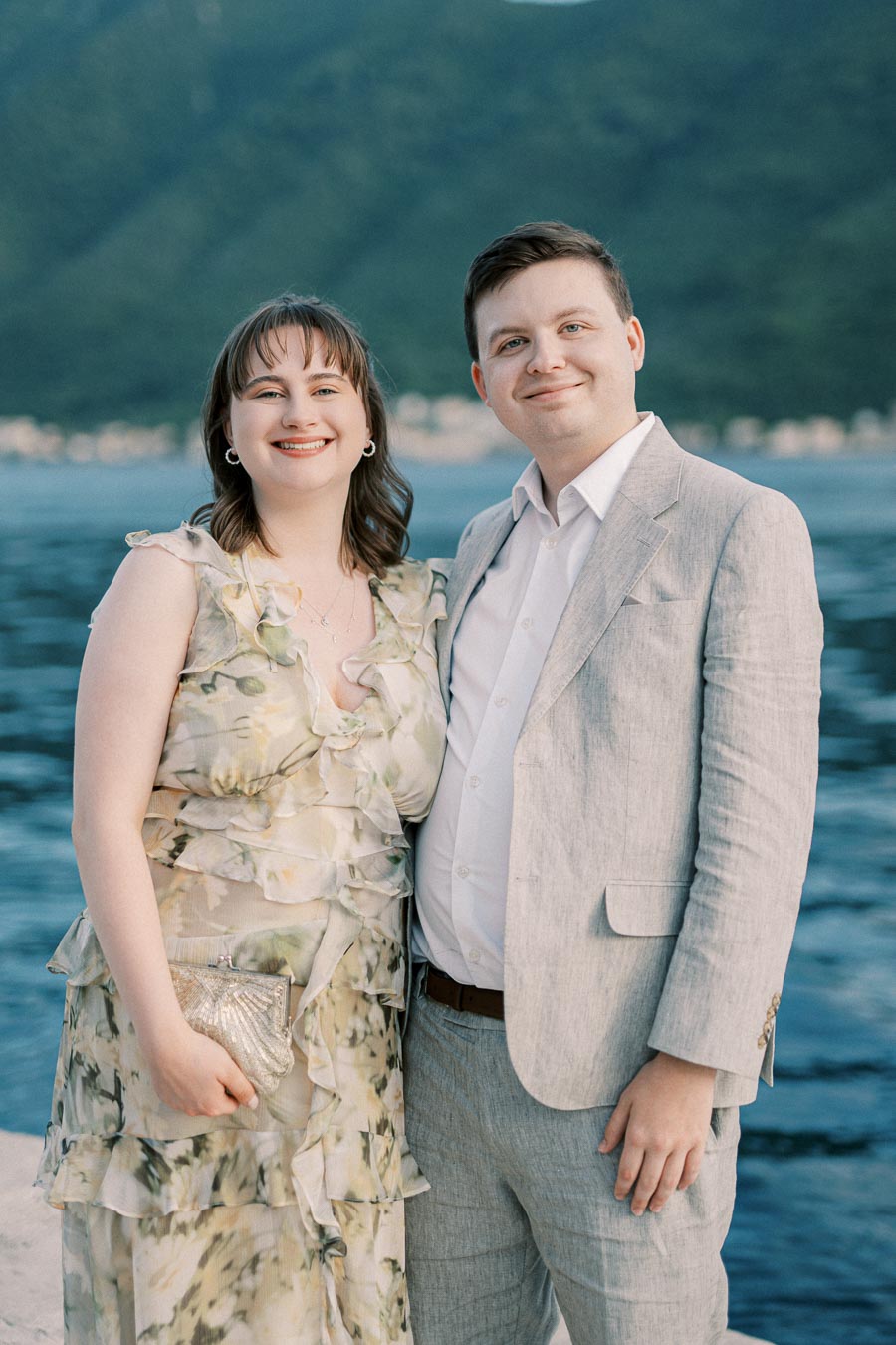 Couple dressed in formal attire standing by a scenic waterfront with mountains in the background.