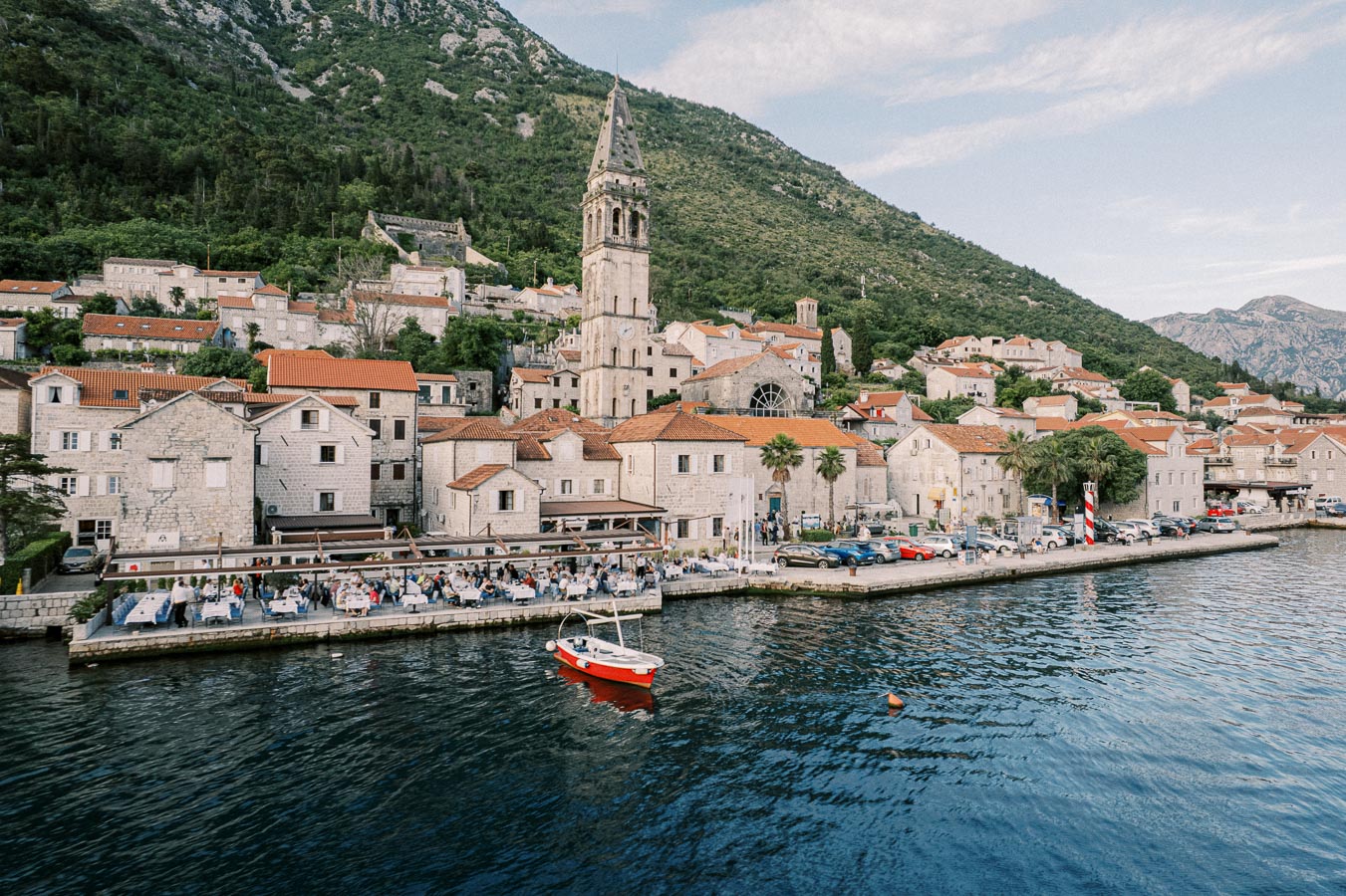 Scenic view of a historic coastal town with stone buildings and a prominent church tower, situated at the base of a lush green mountain, with a vibrant red fishing boat in the foreground on a calm body of water.