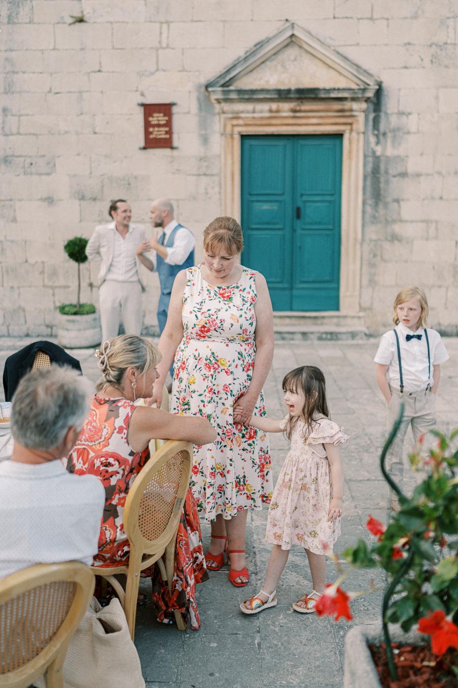 A family gathering in a courtyard with a decorative stone building in the background. A woman in a floral dress holds a little girl's hand, while others sit and converse around a table. A child stands nearby, dressed in formal attire. Red flowers are visible in the foreground.