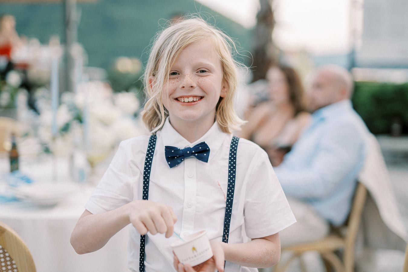 Young child smiling and enjoying ice cream at a formal outdoor event, wearing a white shirt with polka dot suspenders and bow tie.