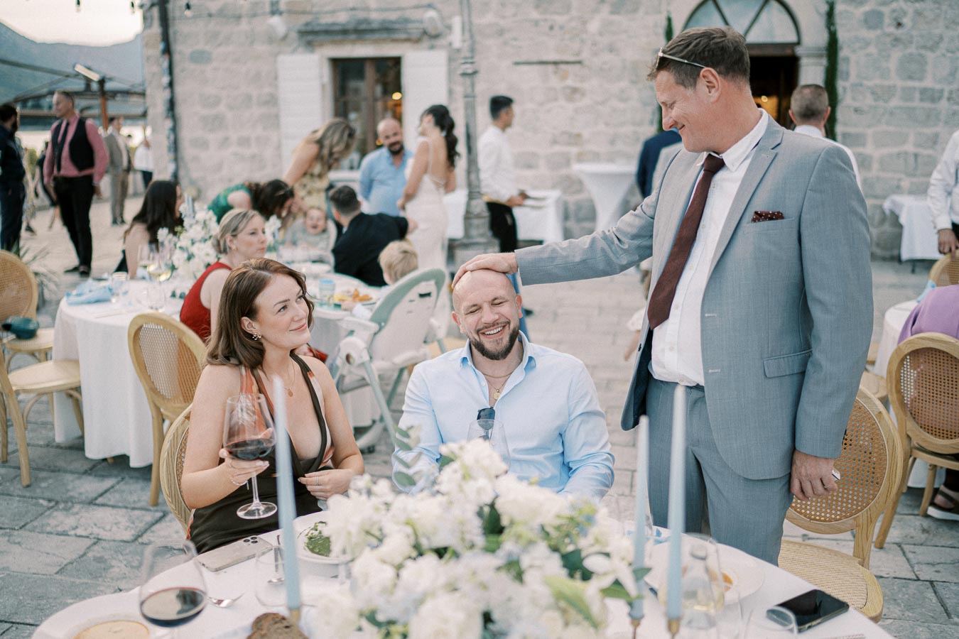 Guests enjoying an outdoor wedding reception; a man in a suit is playfully patting the head of a seated, smiling man with a woman holding a wine glass beside him.