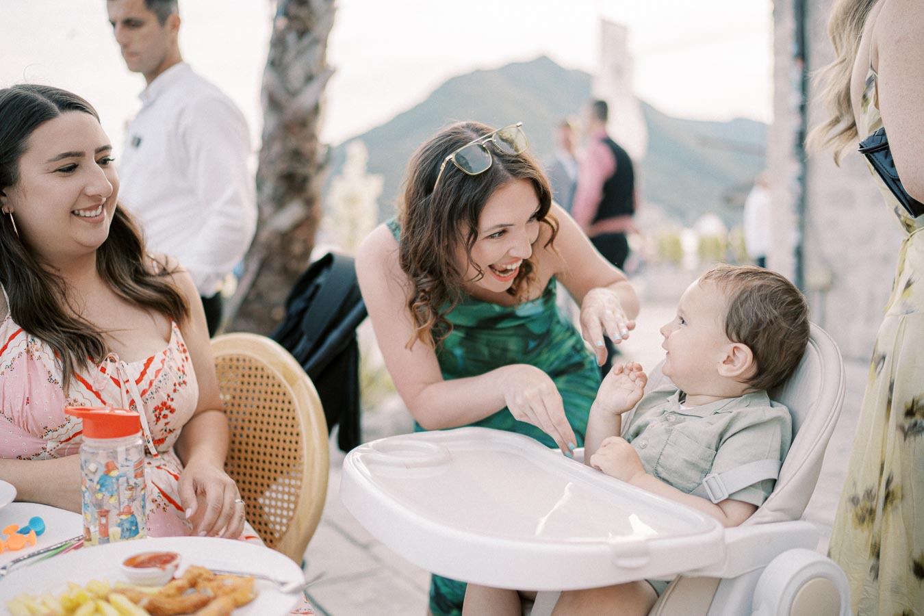A joyful interaction between a smiling woman and a baby in a high chair at an outdoor gathering. The background features a scenic mountain view and a sunny day, creating a warm and cheerful atmosphere. The setting includes a table with food and drinks, indicating a social dining event.
