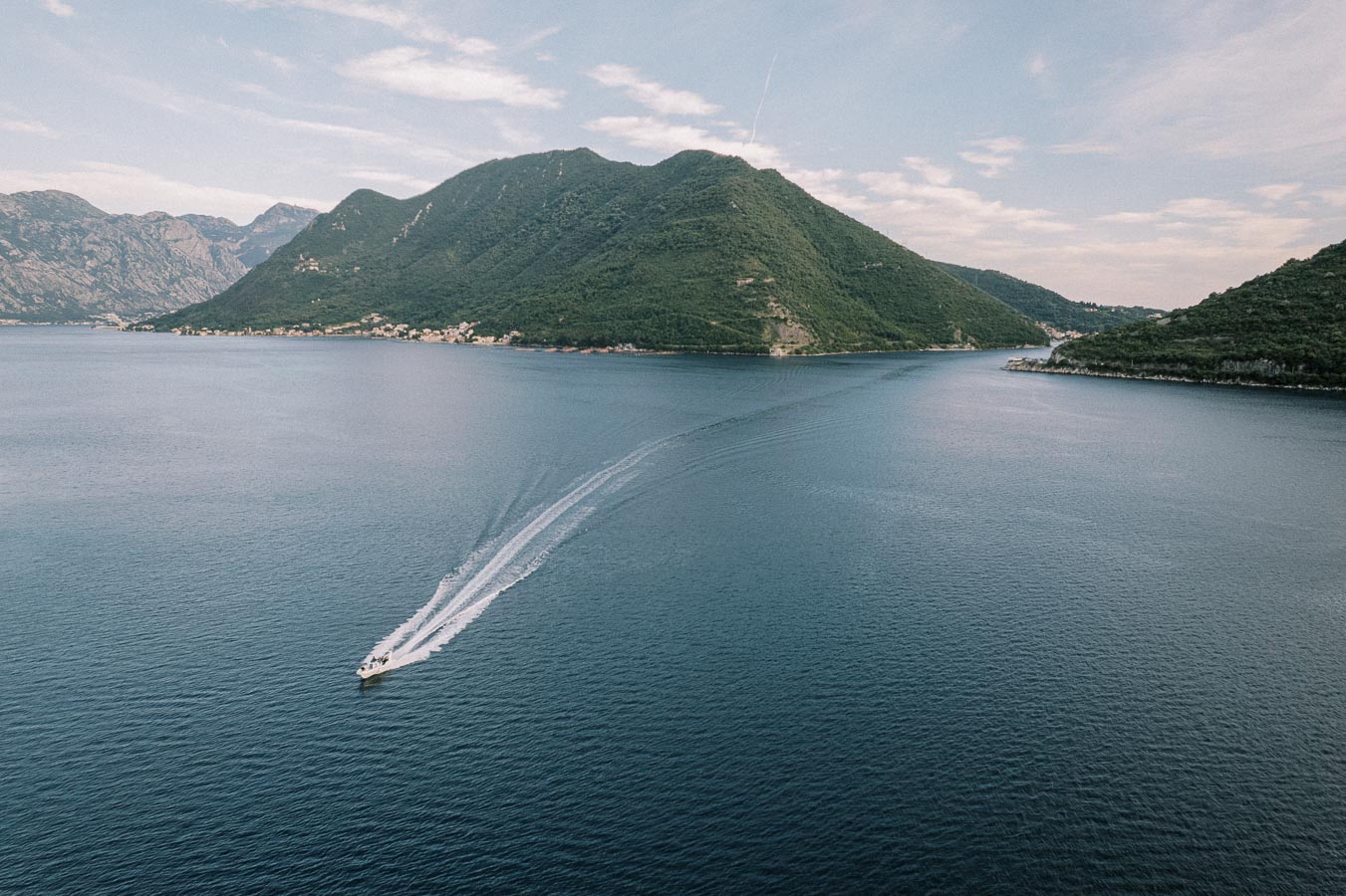 Aerial view of a boat creating a wake on a calm blue bay, framed by lush green mountains under a partly cloudy sky.