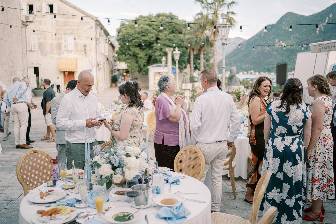 Outdoor wedding reception with guests socializing around decorated tables, set against a scenic backdrop of mountains and a historic building.