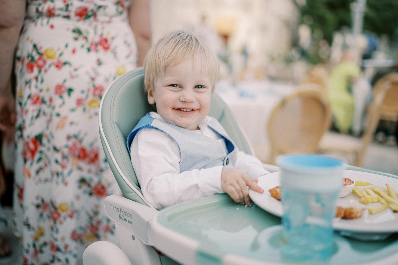 Cute toddler smiling while sitting in a high chair with a plate of fries and chicken, outdoors at a garden party.